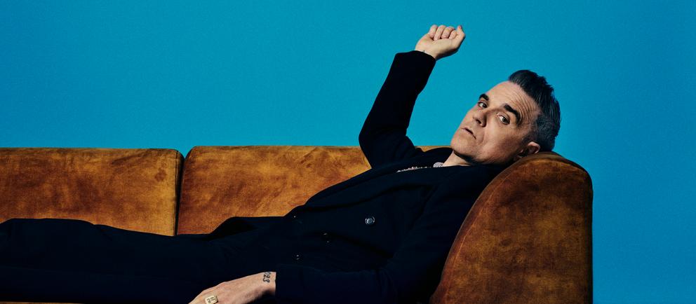 Robbie Williams liegt auf einer braunen Couch und schaut in die Kamera