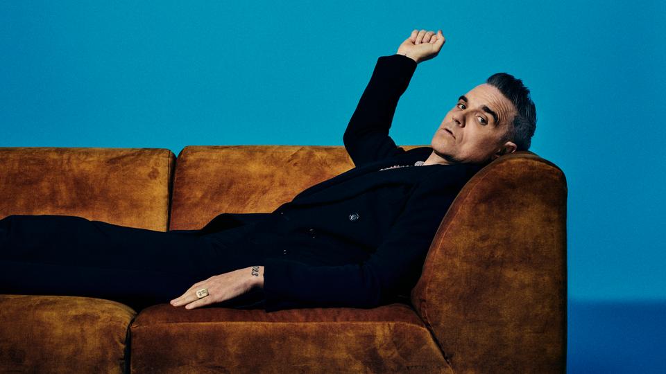 Robbie Williams liegt auf einer braunen Couch und schaut in die Kamera